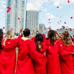 Photo: Shot of a group of graduating students wearing red gowns facing away from the camera and celebrating. Red and white confetti falls from the sky and Stuvi II can be seen in the distance.
