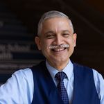 Photo: A man wearing a blue dress shirt and a navy blue vest with a tie leans on a stairwell and smiles for a portrait shot.