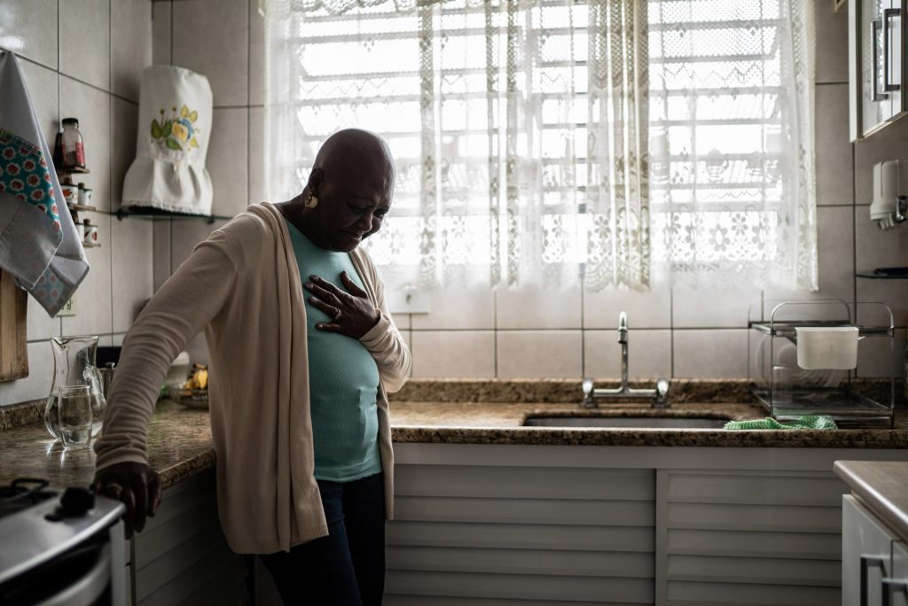 Photo: A senor, Black woman wearing a light blue shirt, tan cardigan, and black pants, has a look of pain on her face as she clutches her chest in her kitchen.