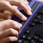 Photo: A set of hands use a computer with a braille computer display and a computer keyboard.