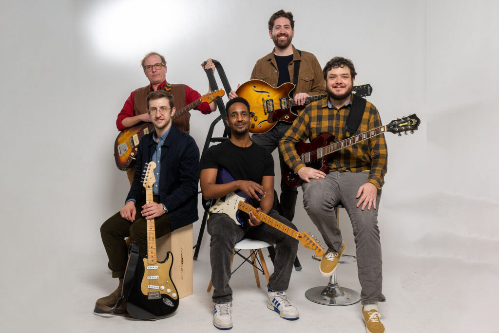 Photo: Group photo of five men sitting and standing. They all hold guitars. From left: Nathaniel Braddock, Brian Barrone, Kumera Zekarias, Erik Broess, and Lance Morrison.
