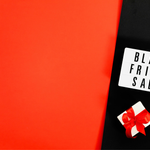 Image: A brightly lit lightbox displaying the words "Black Friday Sale" placed on a black surface next to a small white gift box tied with a red ribbon. The background transitions from solid red on the left to black on the right, creating a striking color contrast.
