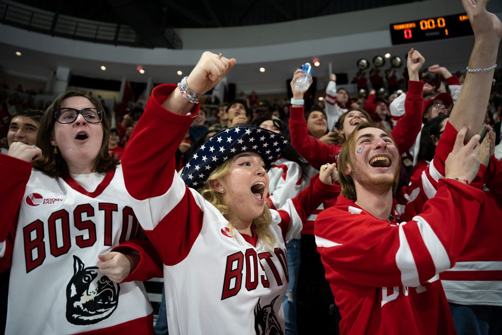 Photo: BU Students wearing large white and red "Boston" ice hockey jerseys cheer and celebrate BU men's hockey in the crowd during a game. Students from left to right: Taelor Anderson(CAS'24), Riley Egan (COM'25), Evan Teplensky(CAS'23).