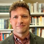 Photo: A man with curly blonde hair posing for a headshot in front of many bookshelves