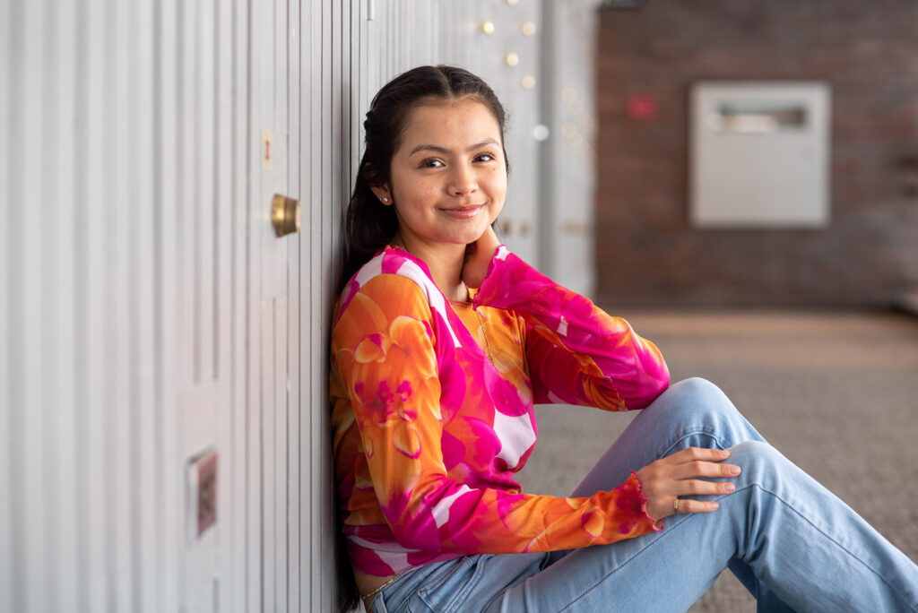 Photo: Kathya Correa Almanza (CAS’25), vice president of Alianza Latina, sits and poses on the floor of a brightly sunlit hallway. A young, Latina woman wearing a pink, orange, and white long-sleeved floral shirt and jeans, poses with one leg bent and arm resting on knee as she smiles for the camera.