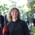 Photo: Megan Ryder, a BU student with curly blonde hair, smiling in front of greenery while holding gardening tools