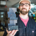 Photo: Micah Sieber, a white man with brown beard and mustache and wearing a tan cap, red-pink shirt, black collared shirt, and black pants, smiles and holds up a small black, model building. The building is in the shape of the BU CDS building, a Jenga-like building with smooth sides.