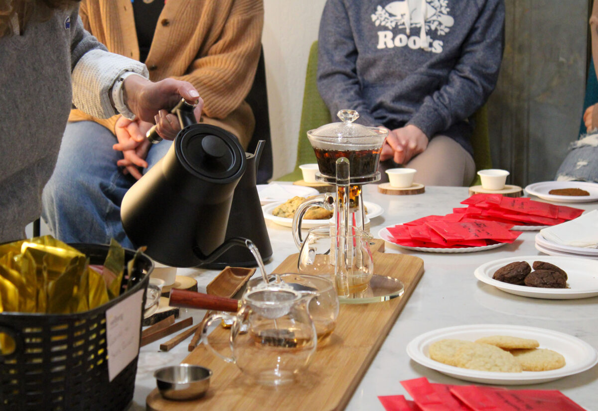 Photo: A table full of various tea accessories is shown. A person to the left is shown carefully pouring hot water into a glass teapot filled with tea leaves, as others around the tables prepare other teas and cups.