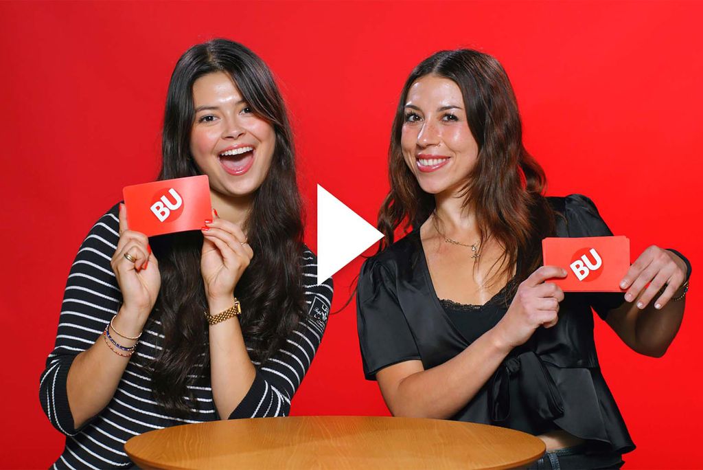 Photo: Two college woman, one wearing stripes on the left and one wearing a black shirt on the right, hold up a red card with BU's logo on the back. They are both smiling wide and are posed in front of a red background. A play button vector is on top of the image.
