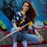 Photo: Sheila del Bosque, a white-presenting woman wearing a yellow, blue, and white collared shirt and jeans, squats and poses with silver flute in left hand. She poses in front of large blue mural.