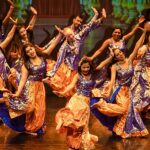 Photo: Colorful Bollywood dancers hit their dance move has the perform on stage. Their costumes consist of sparkly purple tank tops, with sparkly orange bottoms and a navy-blue lining on the pants. Most dancers have their hair down.