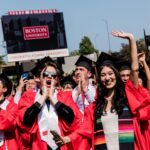 A celebratory scene at Boston University with a group of graduates in red robes and black caps, joyfully raising their hands and capturing photos under a clear blue sky. They are standing in front of a large billboard that reads ‘BOSTON UNIVERSITY CONGRATULATIONS GRADUATES!’ in bold white letters on a red background, marking the festive occasion of their graduation.