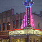 Photo: A brightly lit theater with blues and purples. In art deco style, the sign up top reads "COOLIDGE". Below the sign is a banner for the shows playing. The area is dark as its night tine.