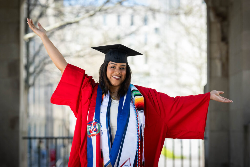 Photo: A college student in a red robe with black mortarboard and blue tassles holds their arms up in celebration