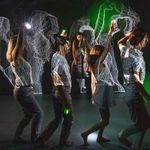 Photo: Four actors walk in a line underneath a wavy laser light display