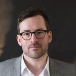 Photo: Formal portrait headshot of a man wearing glasses and a white collared shirt with a grey suit