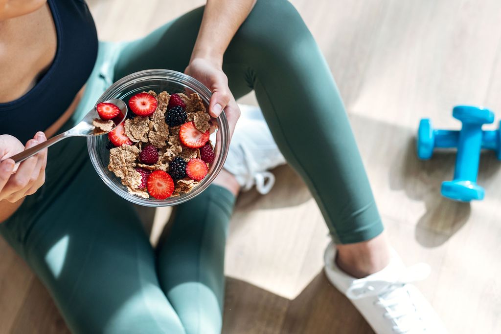 Shot of athletic woman eating a healthy bowl of muesli with fruit sitting on floor in the kitchen at home