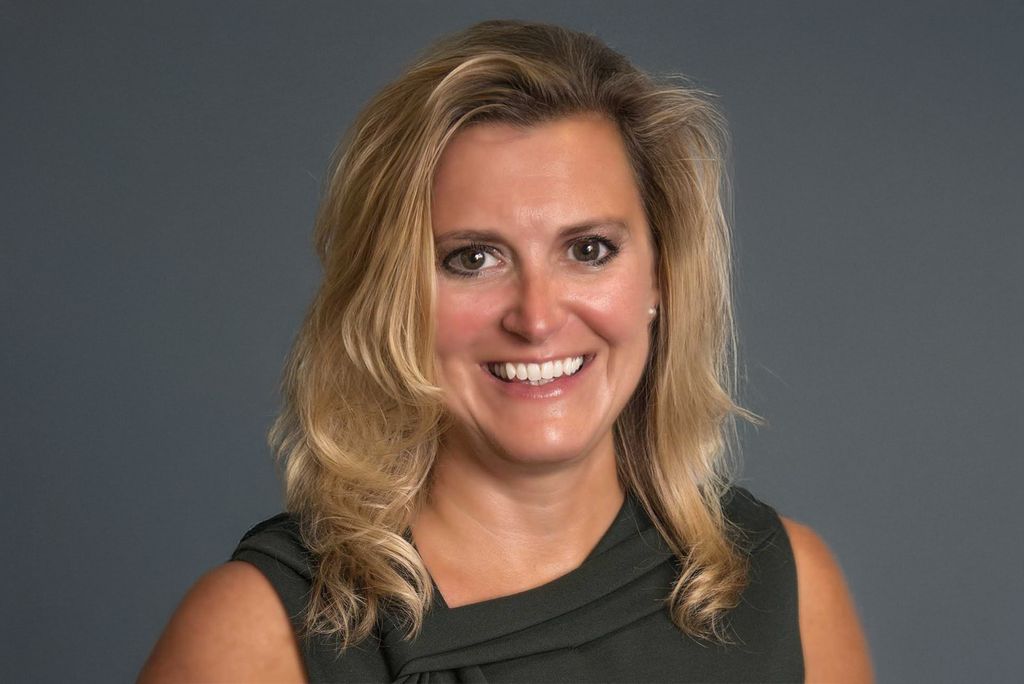 Photo: A white woman with blonde hair in front of a gray background smiles for portrait photo.