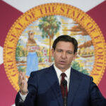 Photo: Ron DeSantis, a white man, stands in front of a banner with Florida's emblem addressing a crowd. He wears a basic navy suit with a red tie.
