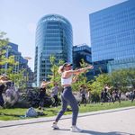 Photo of a women wearing an athletic crop top and leggings punching the air as she speaks into a wireless headset on a bright, sunny day. A group of people in athletic wear can be seen copying her punch as they stand on the green lawn behind her. City buildings can be seen looming in the distance.