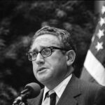 Photo: A black and white photo of Henry Kissinger when he was Secretary of State. He stands at a podium, talking to a crowd in front of a flag of the United States.