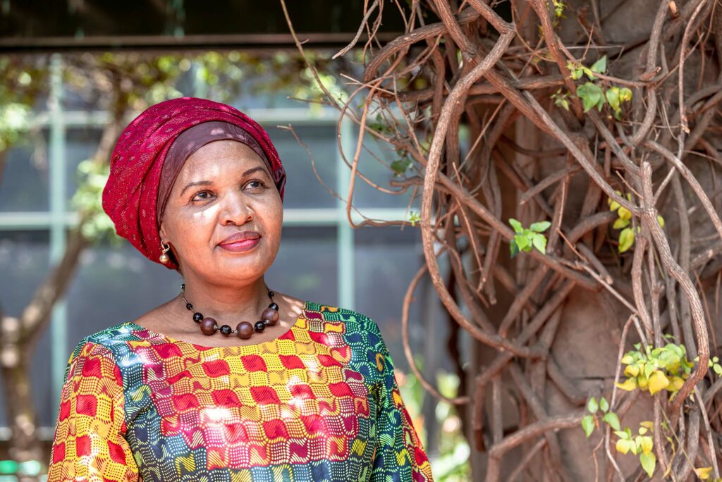 Photo: Esther Kisghu, a Black women, in Kenyan, colorful attire and a red scarf wrapped around her head stands underneath a tree, gazing up at the sky. She is illuminated by the soft light through the tree branches.