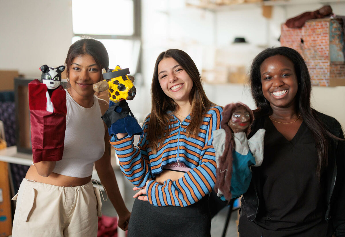 Photo: Three young women hold up puppets on their right hand and smile into the camera. From left, she has short brown hair and wears a monochromatic tan outfit. In the middle, a white woman with long brown hair and a stripped shirt. On the right, a Black woman with shoulder length straight hair. She wears an all black outfit.