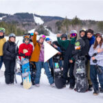 Photo: A panned shot of the entire BU snowboarding team on a snow slope. They are all in their personal gear and warm clothes.