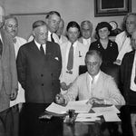 Photo: A black and white photo of various men standing around Franklin D. Roosevelt as he signs the Social Security Bill.