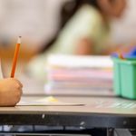 Photo: A close-up of an elementary student writing with a pencil with school supplies on their desk
