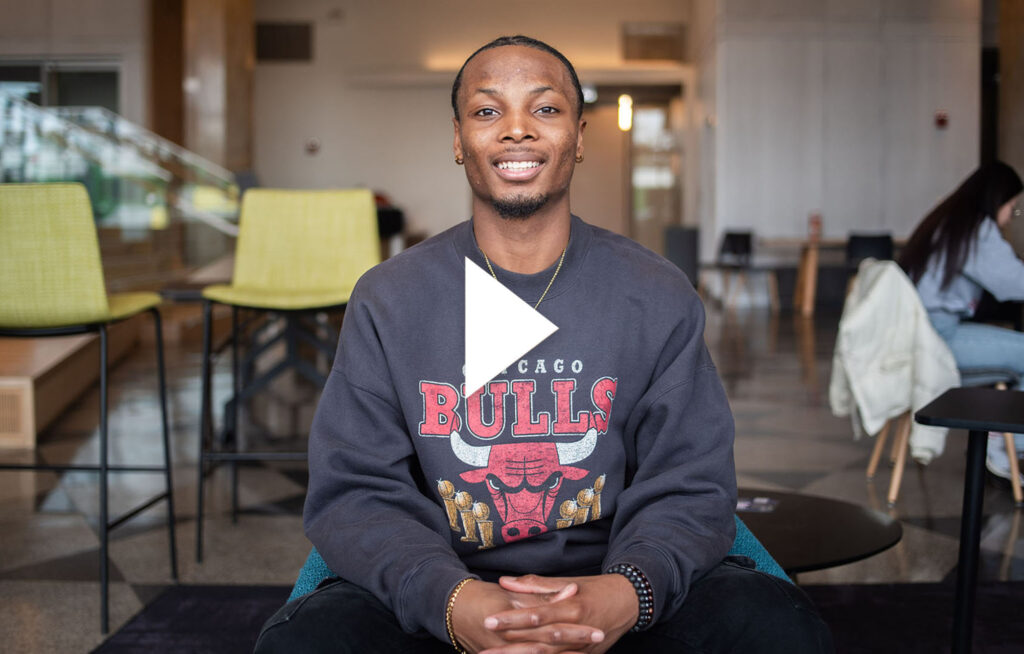 Photo: A young Black man sitting in a chair with a cross around his neck and a Chicago Bulls sweatshirt. He smiles for the photo. Video play overlay