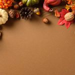 Photo: Brown background with a border of autumnal harvest objects like pumpkins, pinecones, wheat, and acorns