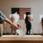 Photo: A person inserts a blank ballot into a wooden ballot box, with a blurred American flag and people in the background.