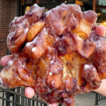 Photo: A hand holding a donut, freshly made, that is bigger than an average plam. Its an old-fashioned donut with a clear glaze. The setting is outside a brick-housed business.