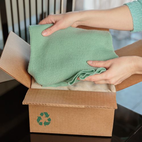 Hands place a green sweater into a cardboard box with a recycling symbol