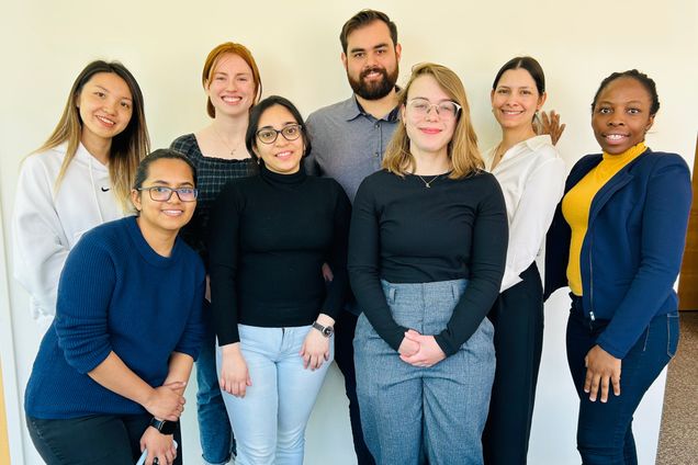 Eight people stand and smile in front of a white wall