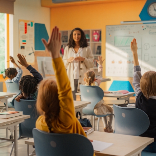 Elementary school classroom with kids raising their hands and a teacher in the front of the room.