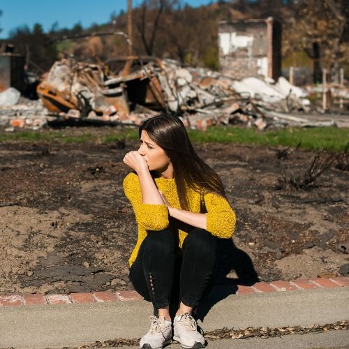 A woman sits in front of a ruined home.