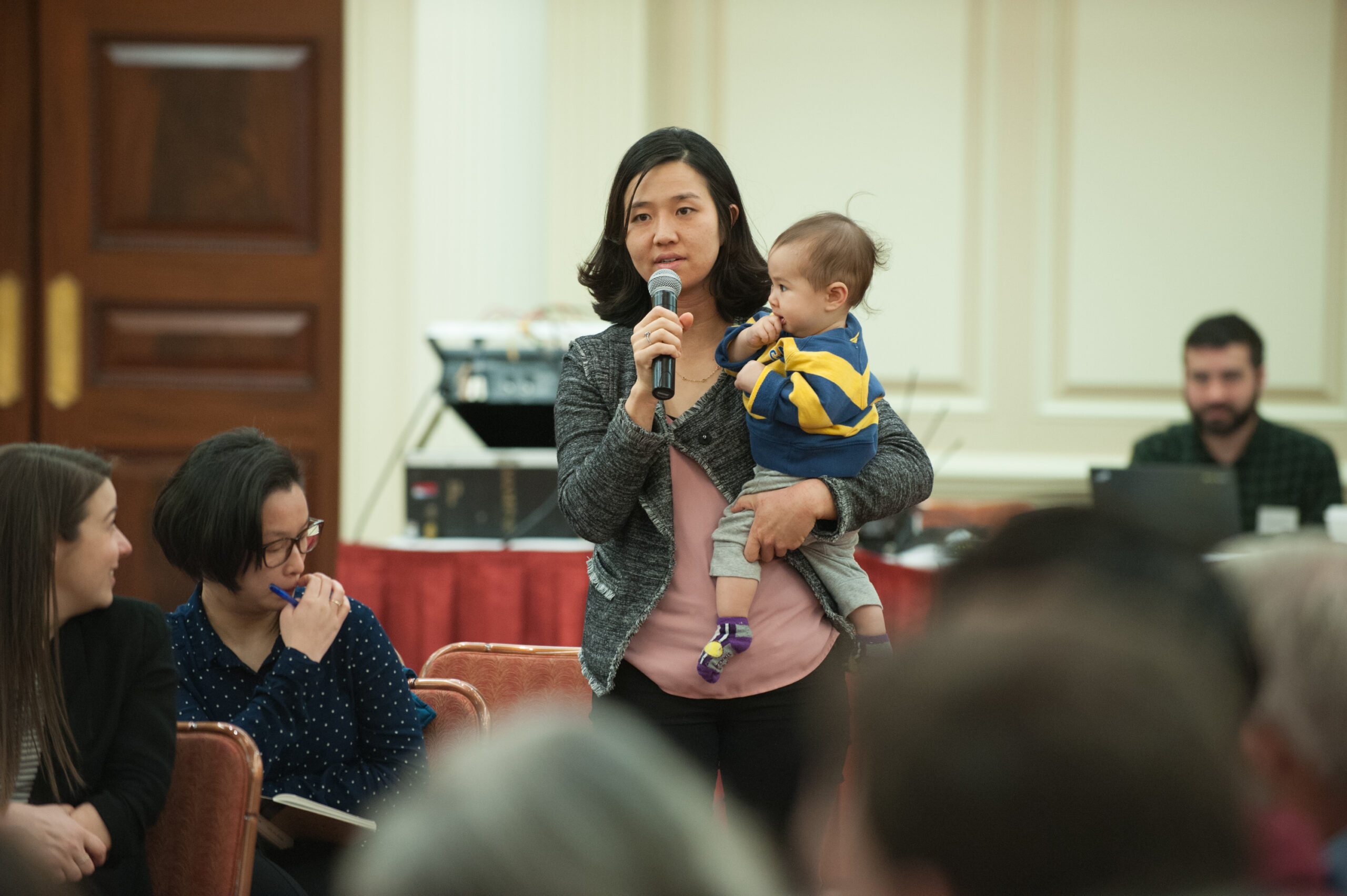 Boston City Councilor At-Large Michelle Wu speaking into a microphone while holding her son, Cass