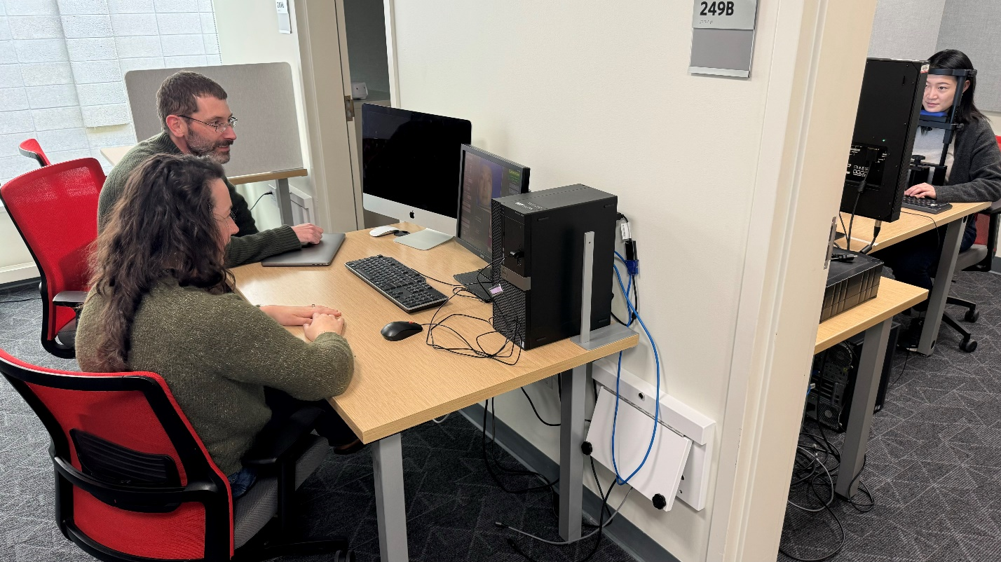 Two people seated at a computer monitor, plus one person at a computer monitor facing opposite