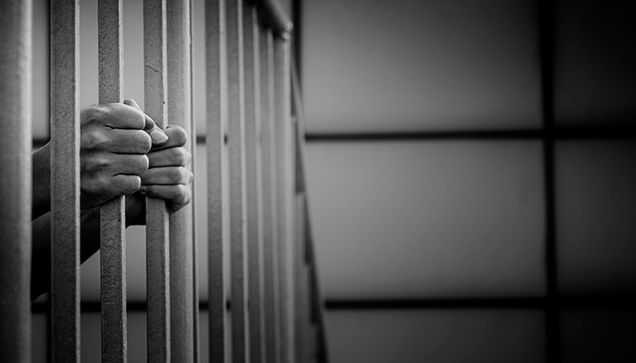 Black and white photo of hands clutching the bars of a prison cell
