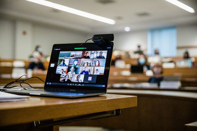 A laptop screen at the front of a classroom displays a Zoom meeting with students learning remotely