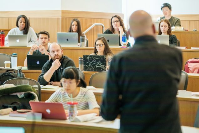 Law professor with his back to the camera teaching students gathered before him in a lecture hall listening intently.