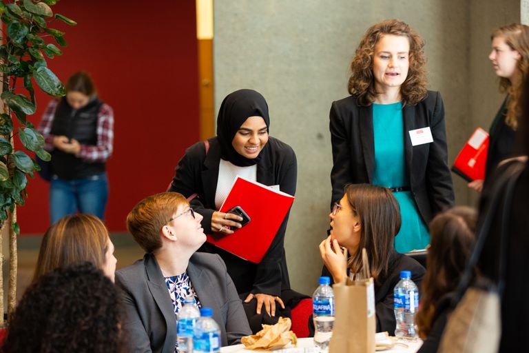Student in hijab holding a red folder leans over desk at LAW MarComm 1L Career Conference to talk to fellow students
