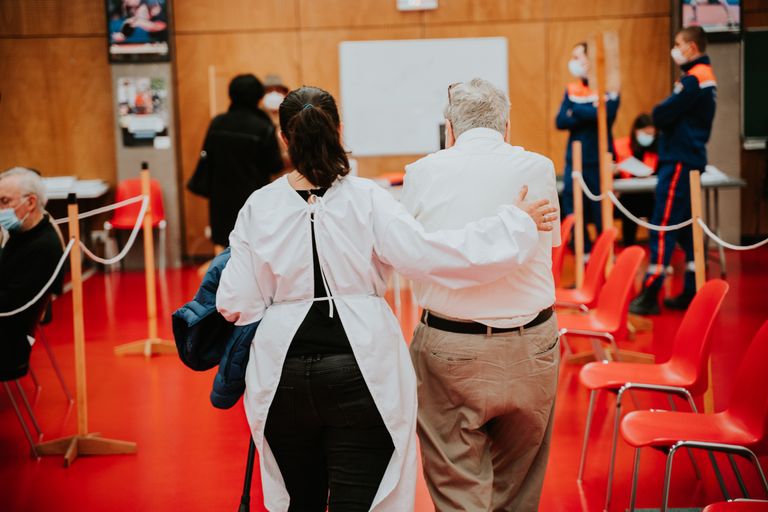 woman in research garb guides an older man down an aisle to get treated