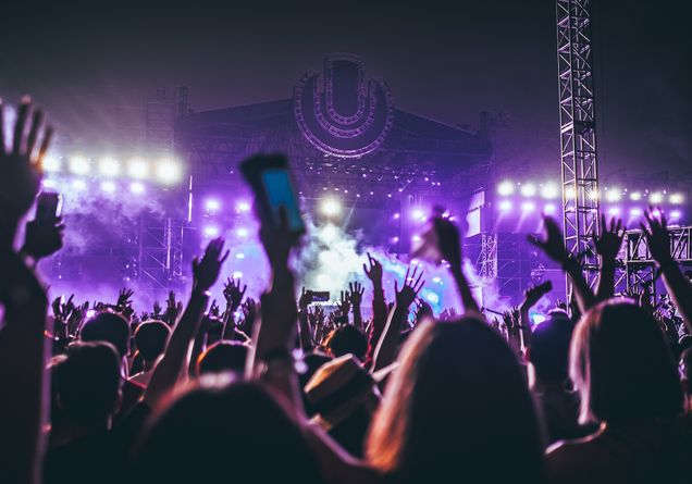 Concert goers hold up phones and wave their hands in front of a large stage with purple lighting