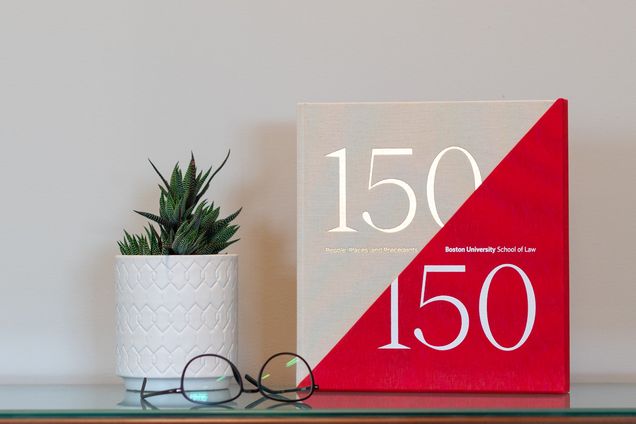 A copy of BU Law's anniversary book, 150/150: People, Places, and Precedents, sits on a table with a plant and a pair of glasses