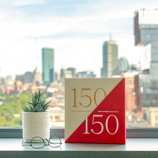 150/150 book on a windowsill alongside a plant and glasses with the Boston skyline in the background.