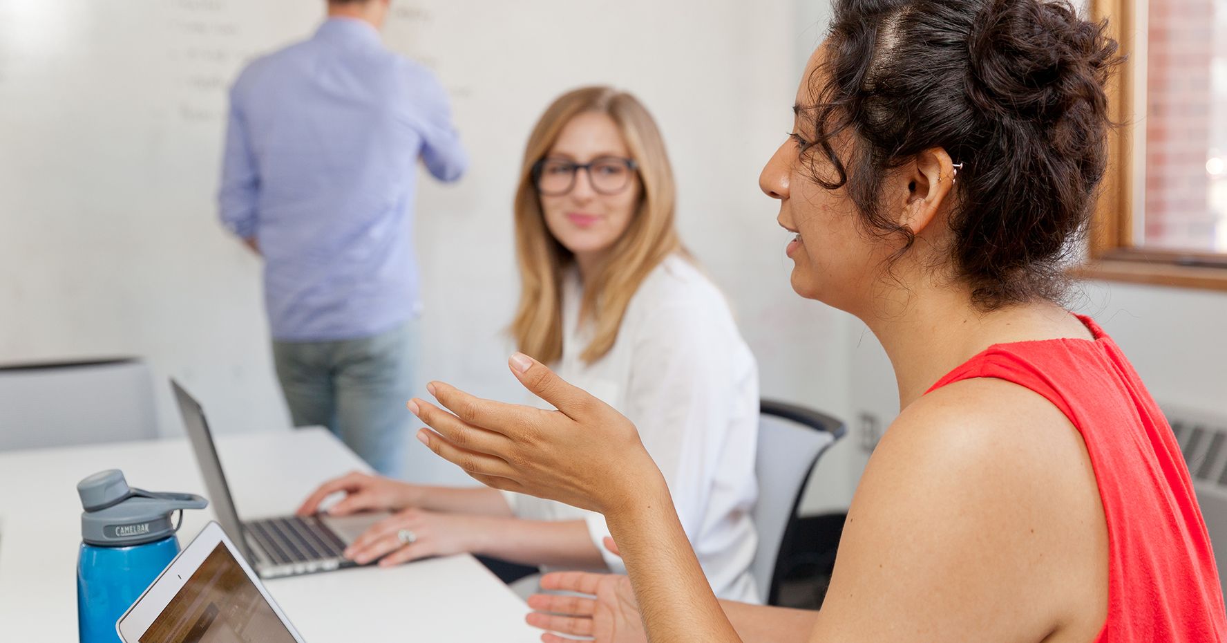 Two female students are engaged in conversation while a professor writes on a whiteboard in the background.