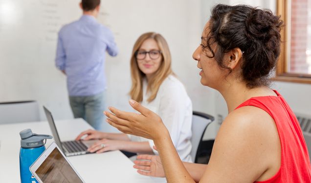 Two female students are engaged in conversation while a professor writes on a whiteboard in the background.
